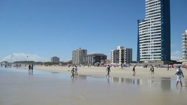 A tall waterfront tower rises beside people in shallow water.
