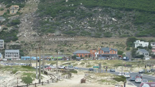 Cars follow a coastal road beneath houses on a green slope.
