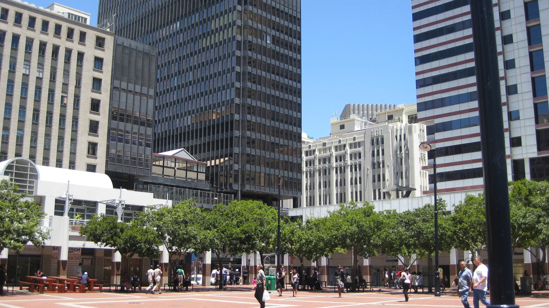 People cross a square bordered by trees and tall buildings.