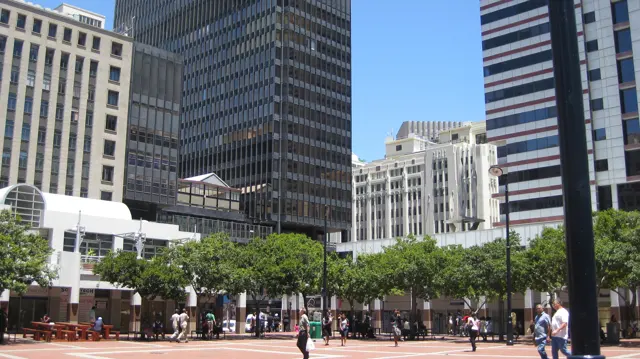 People cross a square bordered by trees and tall buildings.