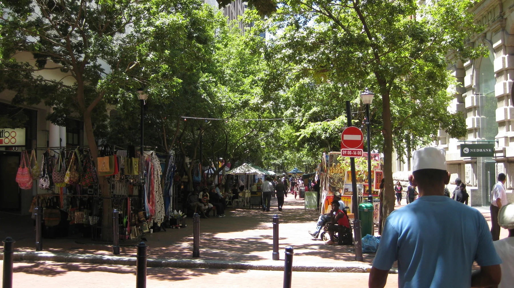 People walk past colorful stalls along a tree-shaded street.