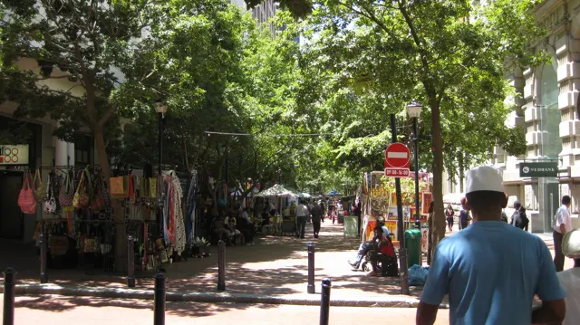 People walk past colorful stalls along a tree-shaded street.