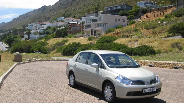 A pale car is parked beneath houses on a green slope.