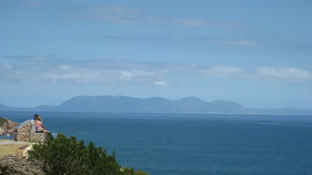A person at a rocky lookout faces a distant mountain across water.