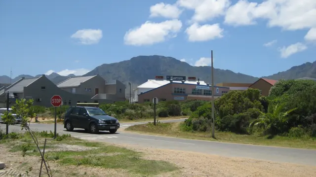 A car passes low pale-roofed buildings beneath a mountain.