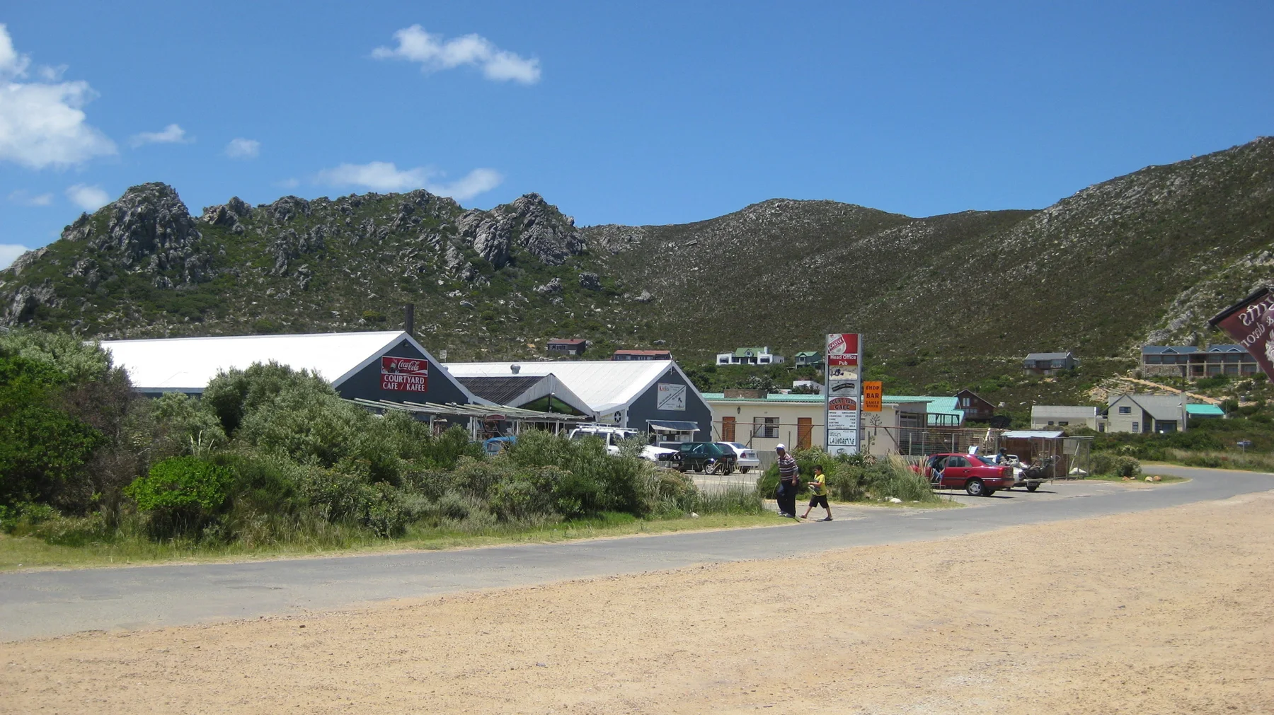 Small shops stand beside a broad roadside below rocky hills.