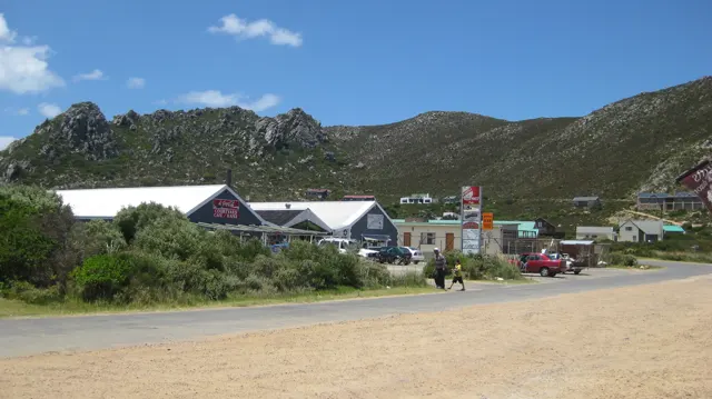 Small shops stand beside a broad roadside below rocky hills.