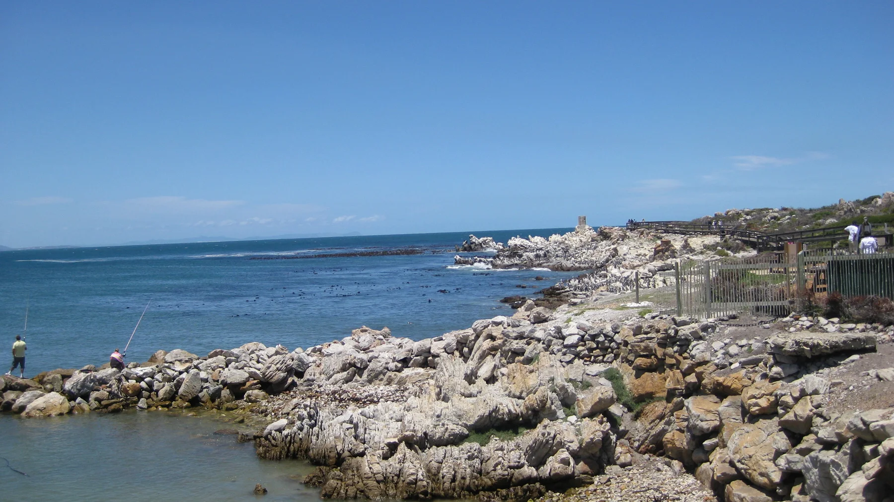 Pale rocks line blue water beside a fenced coastal path.