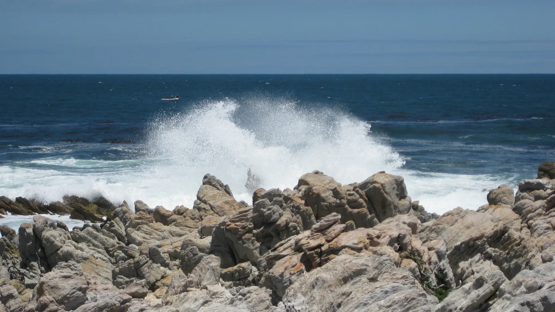 A wave bursts into white spray behind jagged rocks.