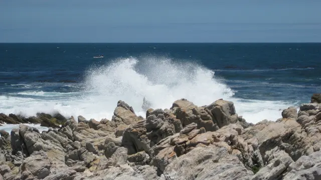 A wave bursts into white spray behind jagged rocks.