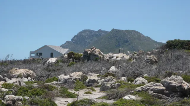 A small pale house stands beyond rocks beneath a green hill.