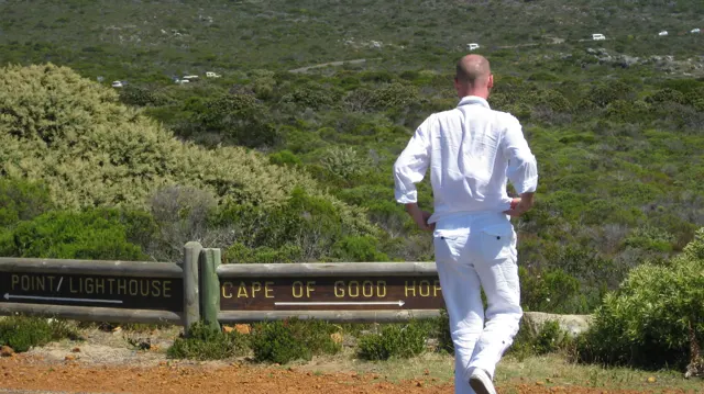 A person dressed in white stands beside wooden Cape direction signs.