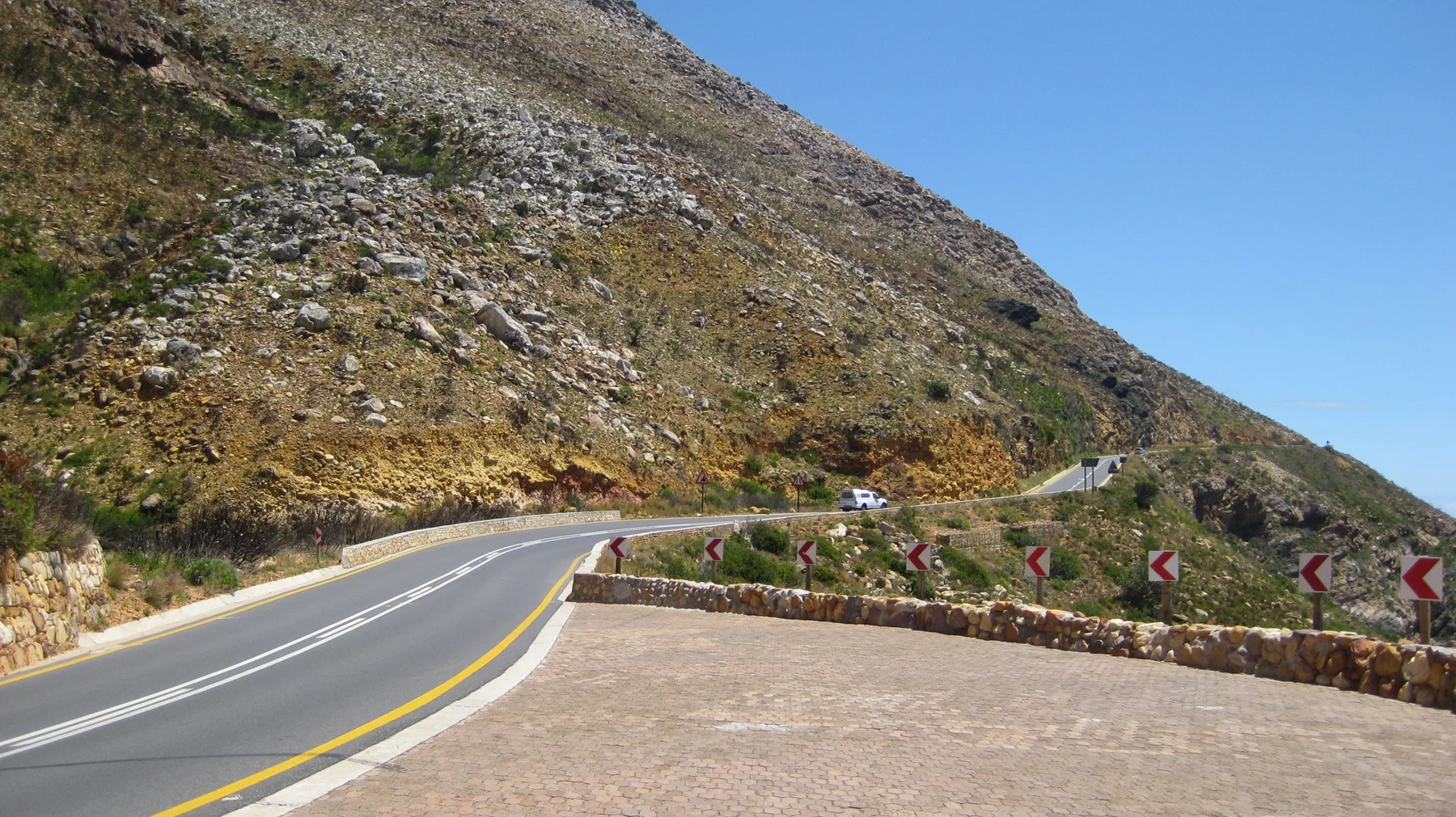 A road bends around a rocky slope beside a stone lookout wall.
