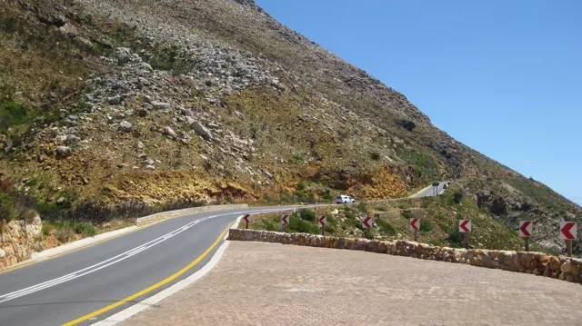 A road bends around a rocky slope beside a stone lookout wall.