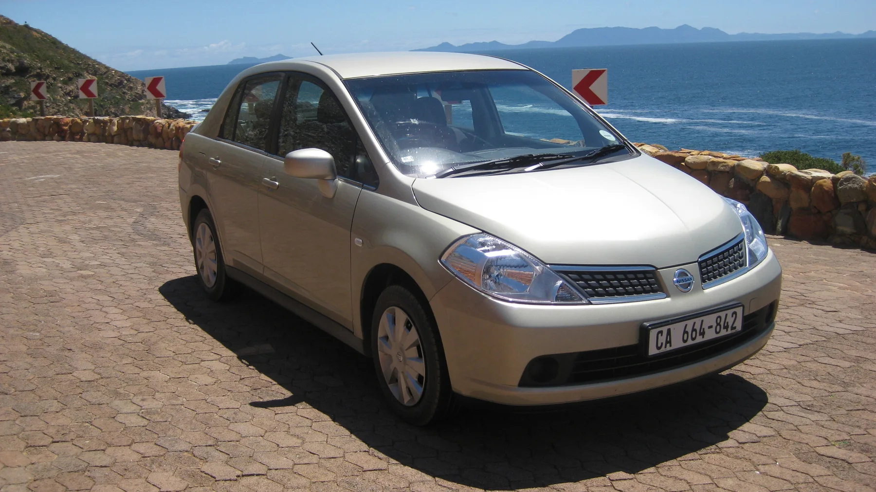 A pale car stands at a paved lookout above blue sea.