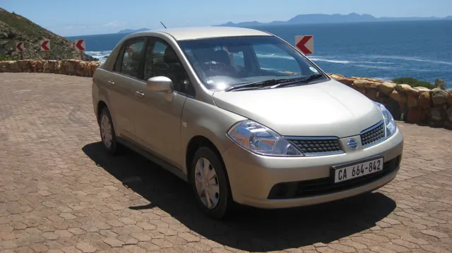A pale car stands at a paved lookout above blue sea.