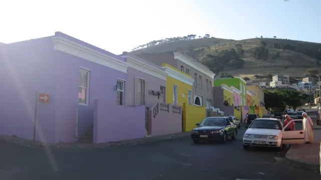 Lavender and yellow houses border parked cars on a sloping street.
