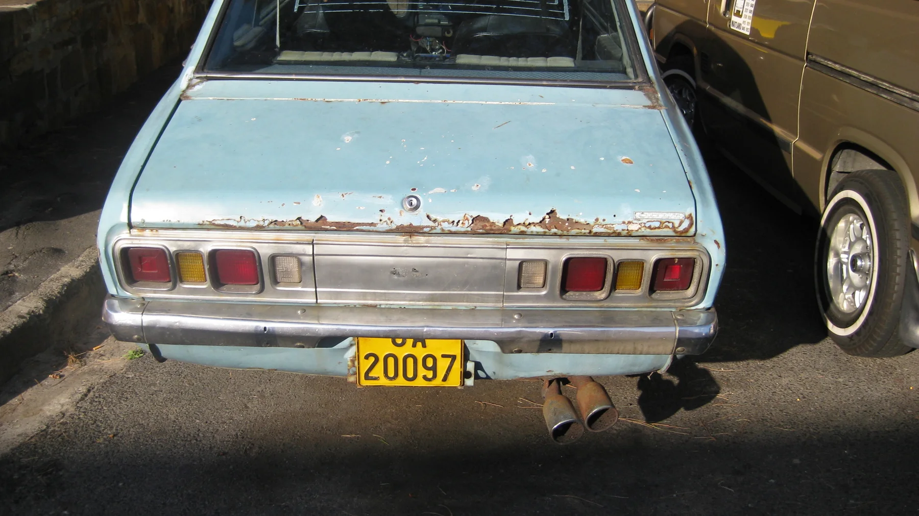 The rear of a weathered pale-blue car shows rust and rectangular lights.