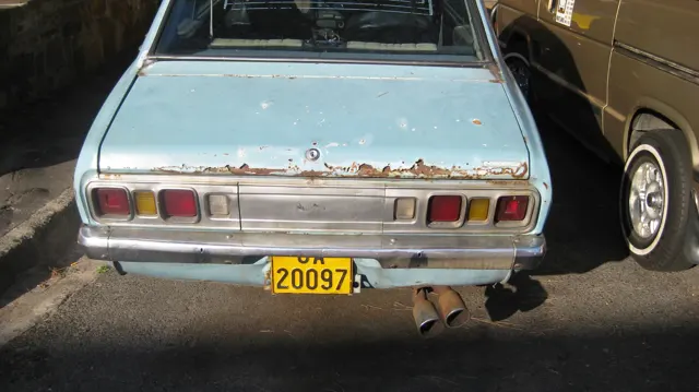 The rear of a weathered pale-blue car shows rust and rectangular lights.