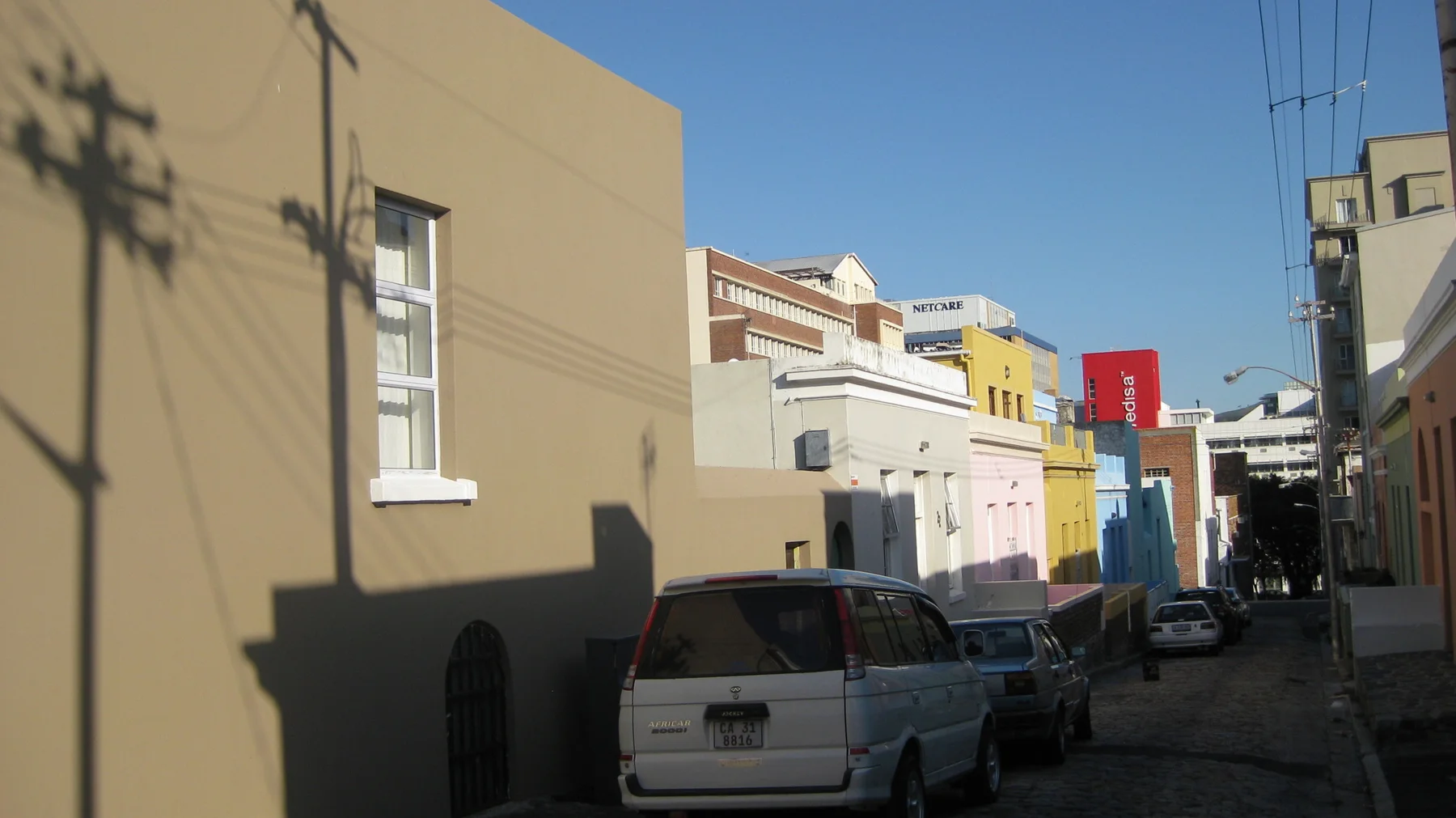 Utility shadows cross a pale facade beside parked cars.