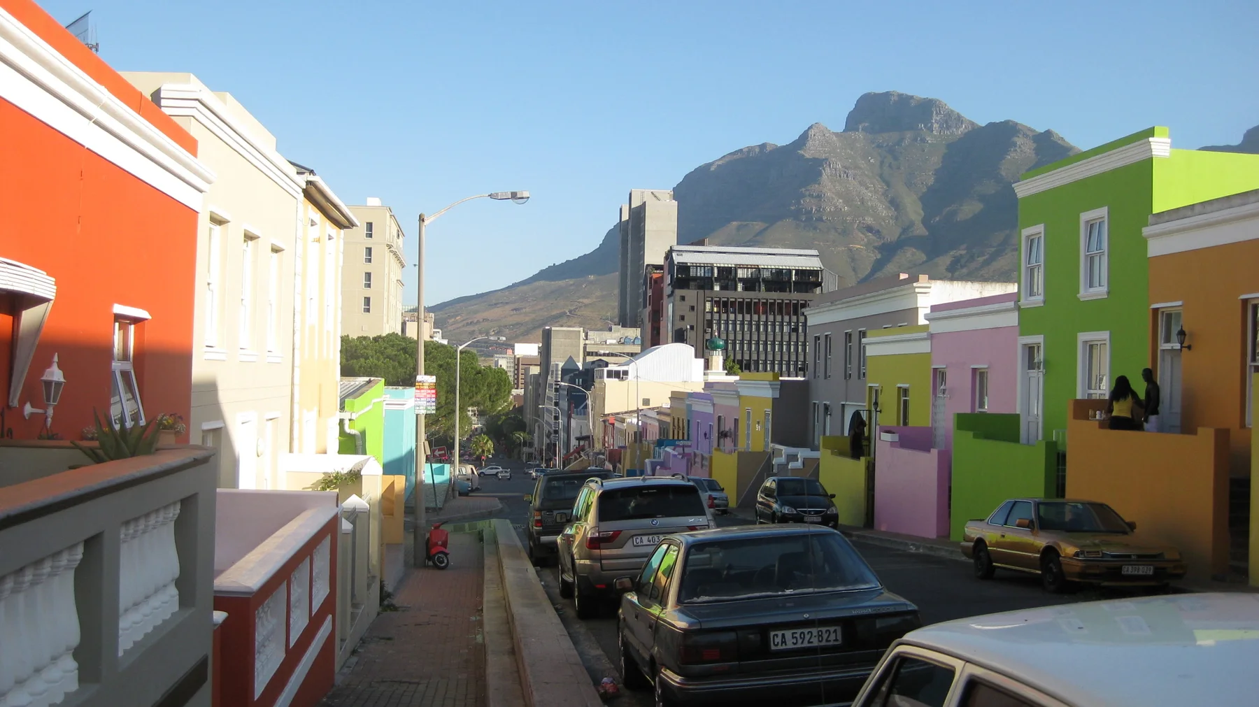 Parked cars fill a narrow street between brightly painted houses.