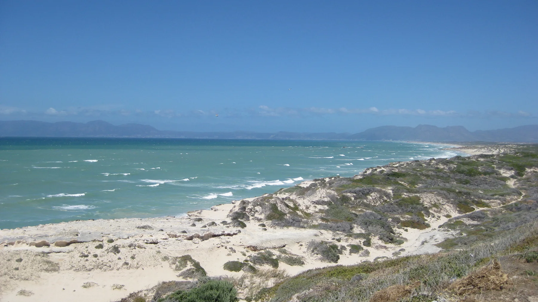 Pale dunes and coastal scrub border a long stretch of turquoise water and white surf.