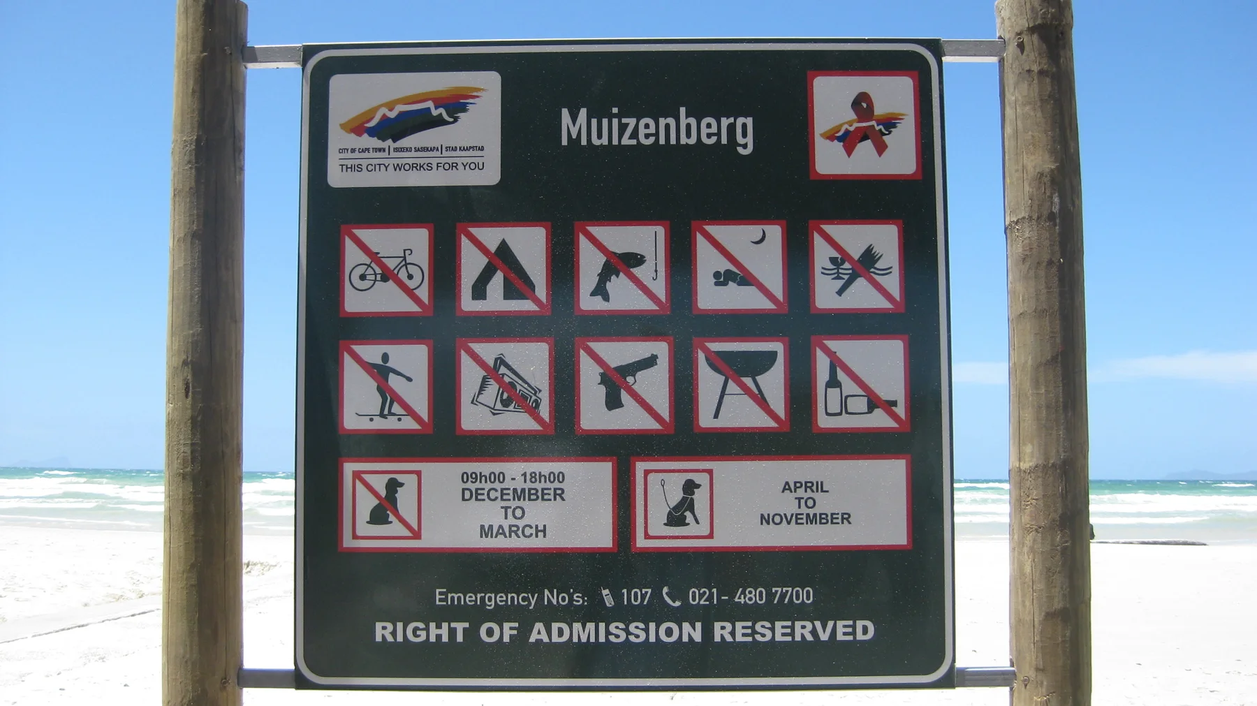A Muizenberg sign displays beach activity symbols beside wooden posts and the water.