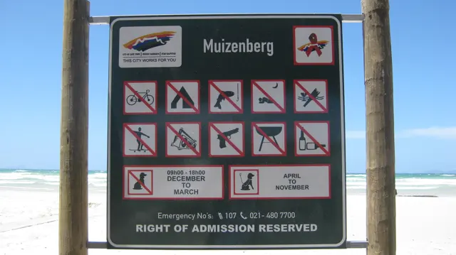 A Muizenberg sign displays beach activity symbols beside wooden posts and the water.