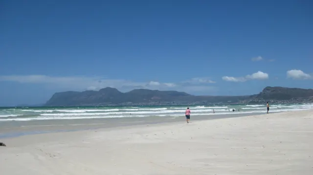 A few distant visitors walk along a broad white beach beneath coastal mountains.