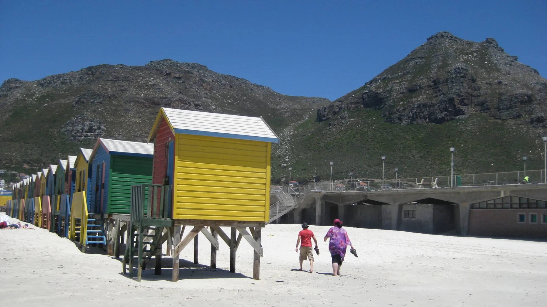Green and yellow beach huts stand on stilts beside people walking across the sand.