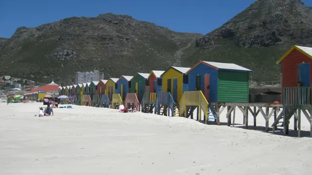 A row of colourful beach huts stretches across the sand with mountains behind.