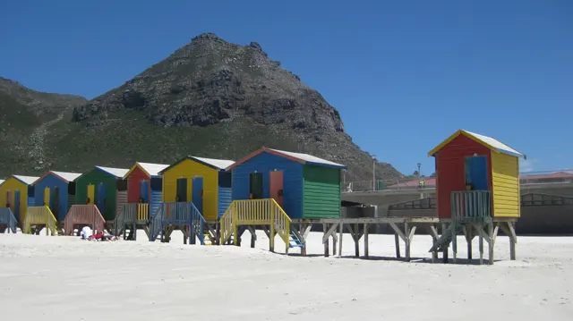 Colourful beach huts and yellow steps stand beneath a pointed mountain peak.
