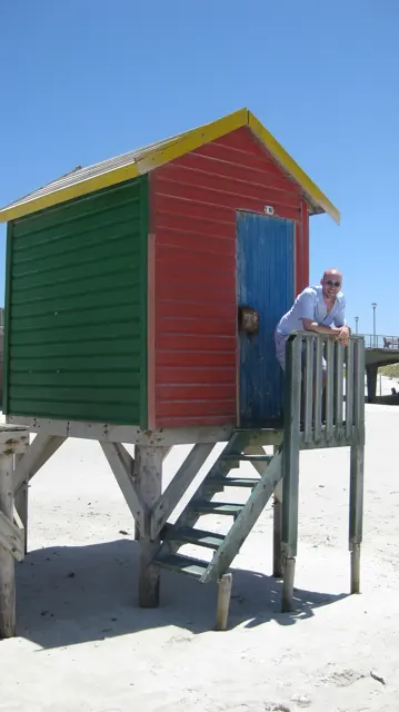 A person stands beside the steps of a green beach hut with bright red panels.