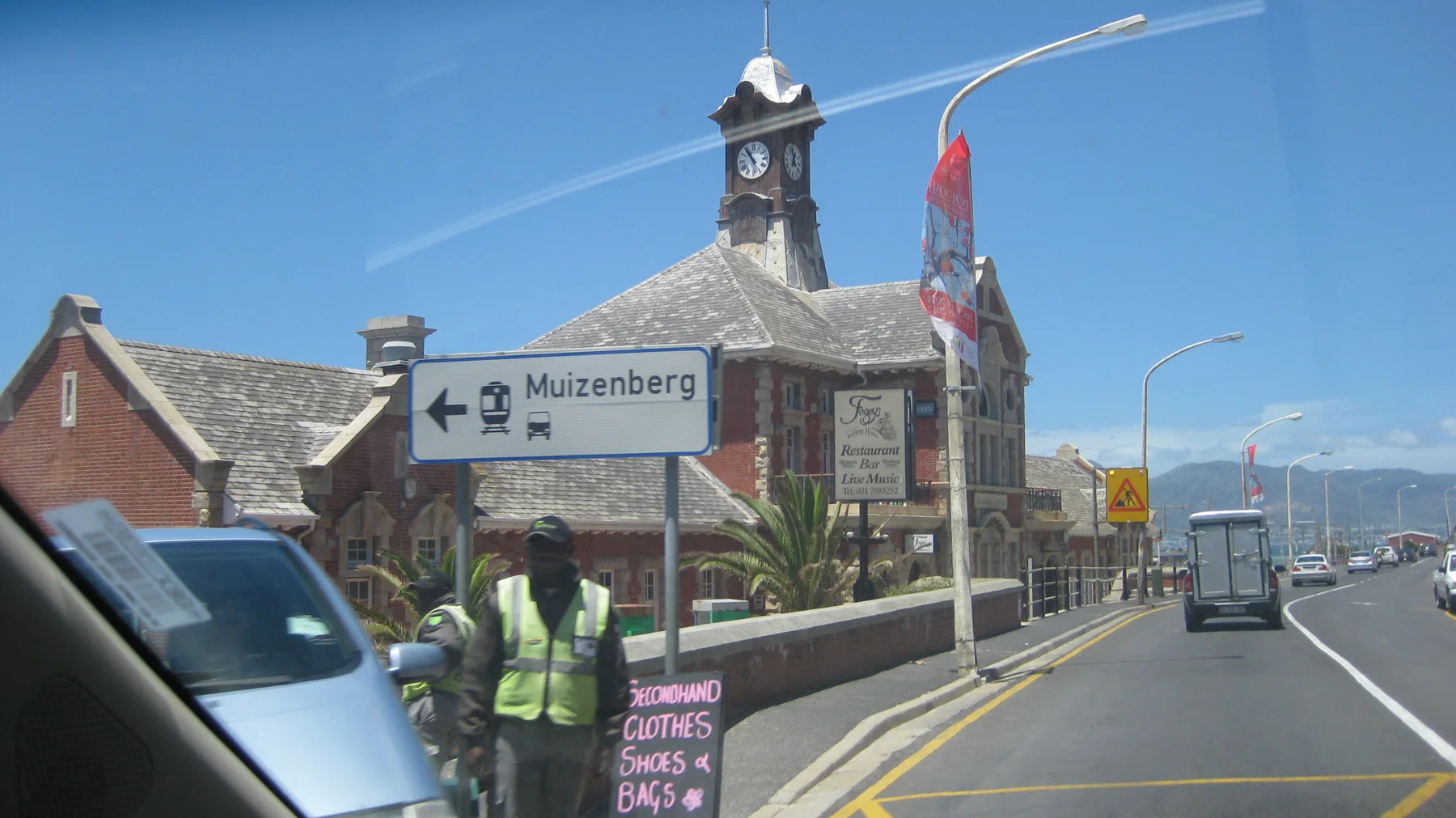 A Muizenberg direction sign stands near a stone building with a clock tower.