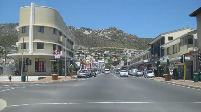 A pale yellow corner building overlooks an intersection beneath a hillside dotted with houses.