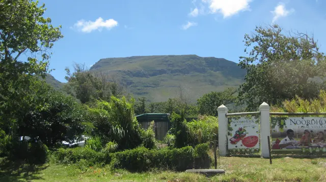 Trees and signs mark a garden entrance with a green hillside beyond.