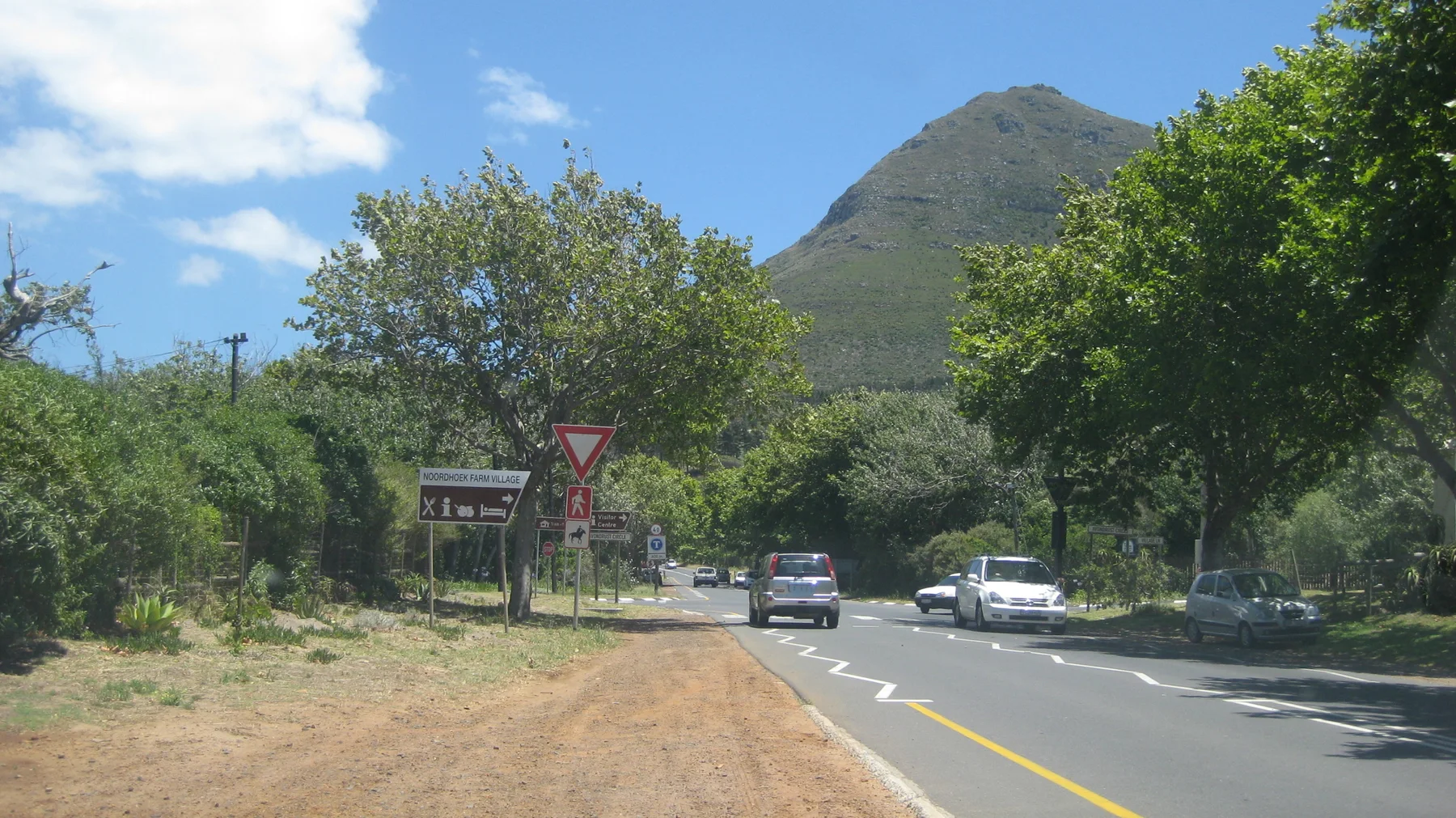 Cars follow a road bordered by trees with a hill rising in the distance.