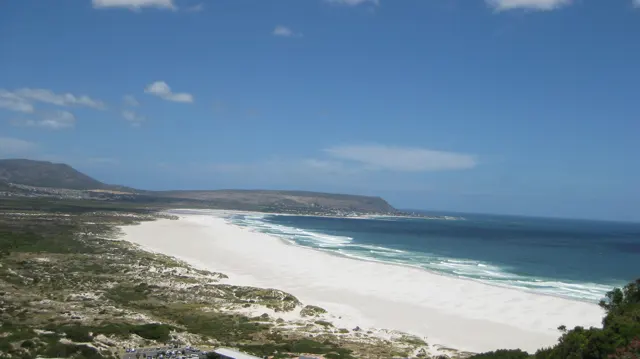 An elevated view follows a long white beach between dark blue water and green dunes.
