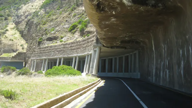 A coastal road passes beneath a rock overhang supported by concrete pillars.