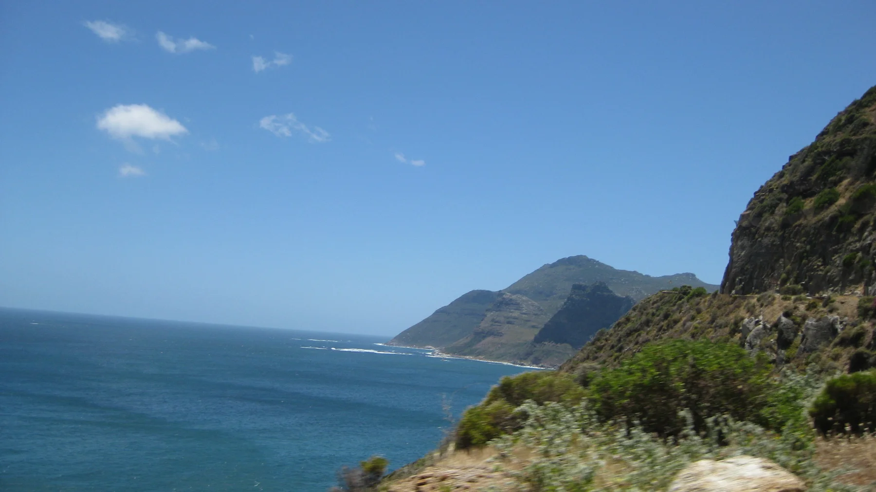 A road curves along a green rocky cliff above blue water and a distant headland.