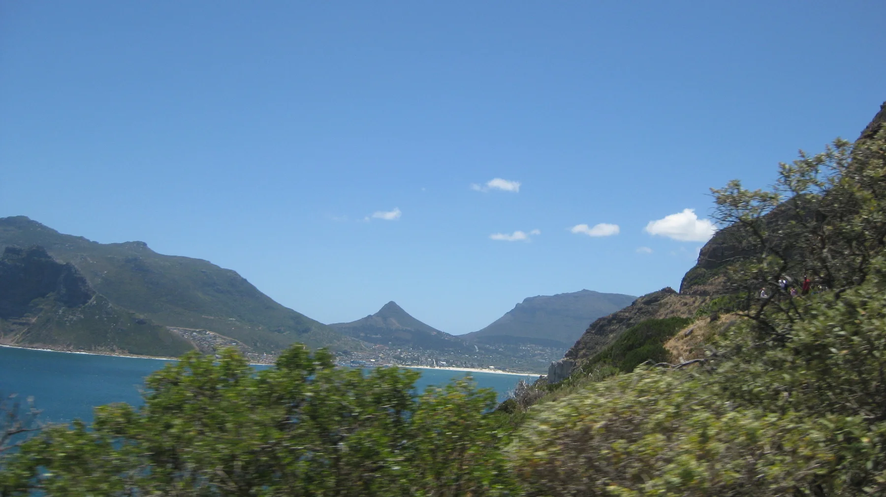Foreground trees frame a bay, a winding cliff road and a pointed mountain beyond.