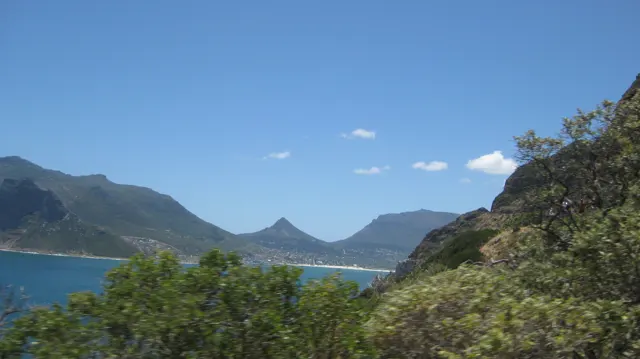 Foreground trees frame a bay, a winding cliff road and a pointed mountain beyond.