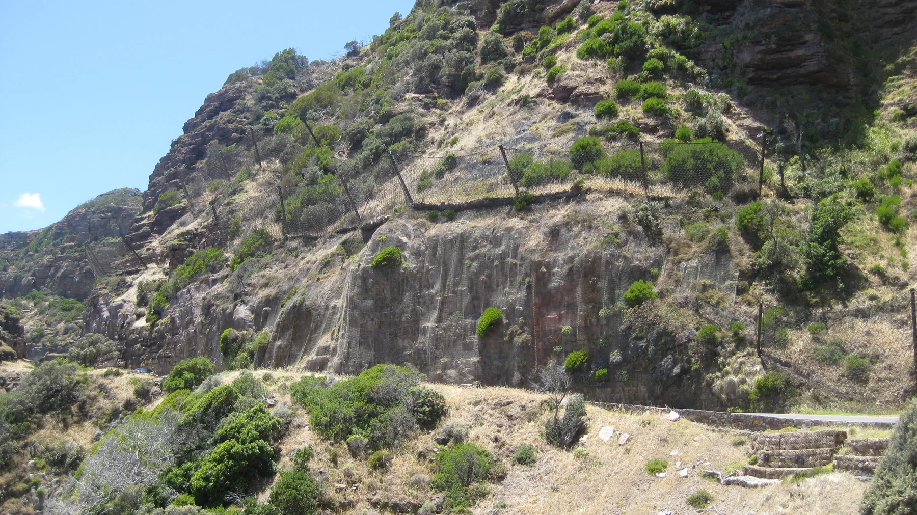 A reinforced road cuts horizontally across a steep green and rocky mountainside.