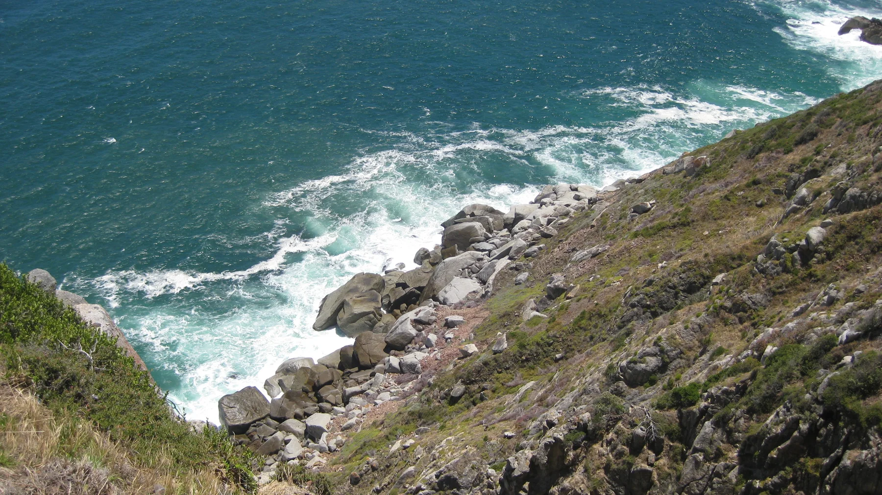 White waves curl around rocks beneath a steep coastal slope.