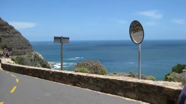 A convex traffic mirror and roadside signs stand above a stone wall beside the sea.