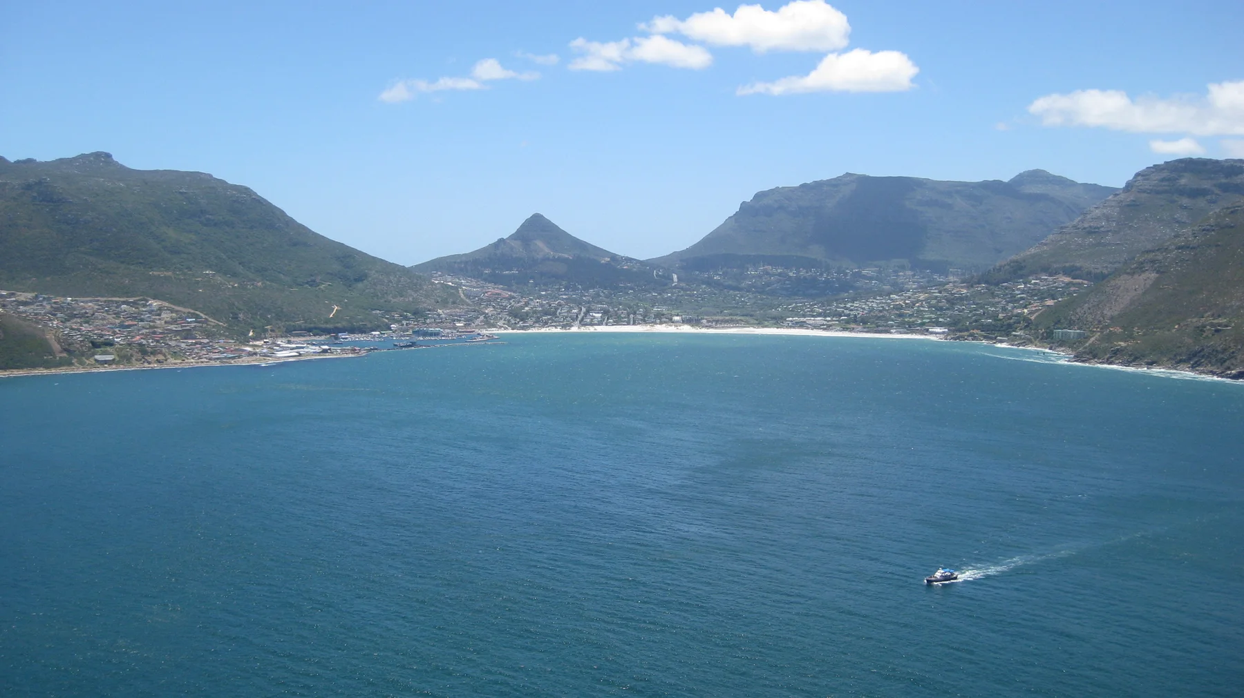A small motorboat leaves a white wake across a broad bay backed by mountains and buildings.