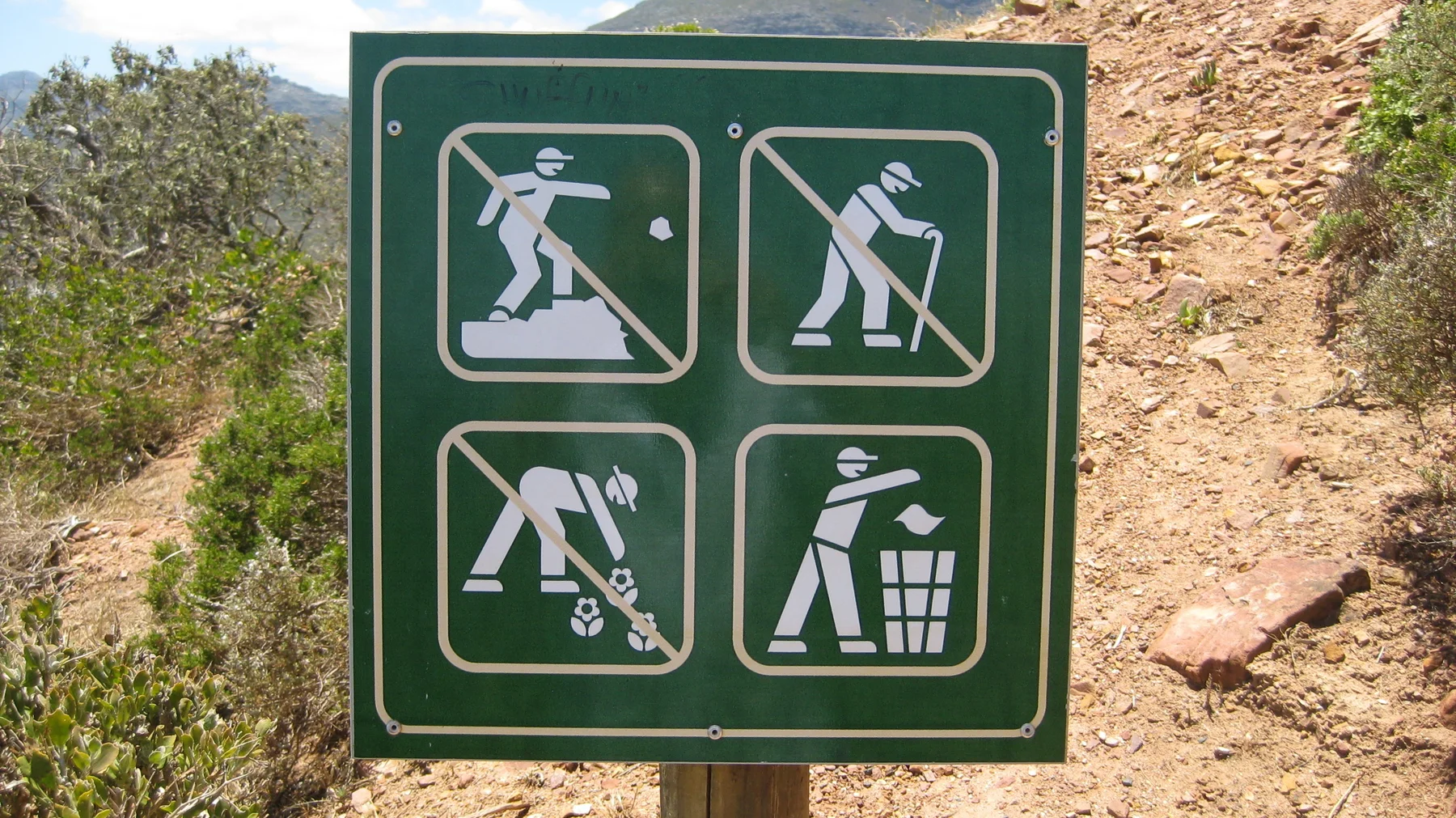 A green outdoor sign displays visitor-care pictograms beside reddish rock.