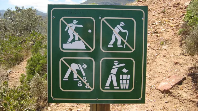 A green outdoor sign displays visitor-care pictograms beside reddish rock.