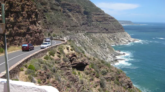 Cars follow a narrow road beneath a high cliff with white waves breaking below.