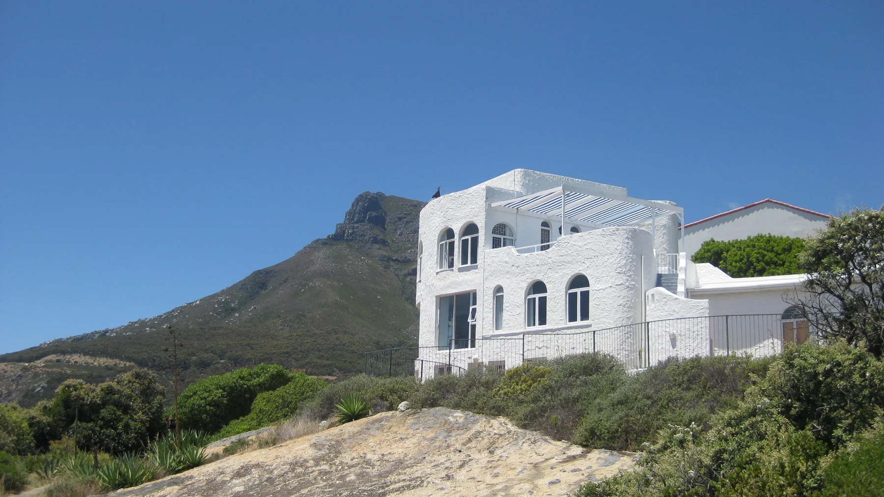 A large white house with arched windows stands on a rocky hillside beneath blue sky.